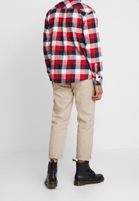 Camisa de franela a cuadros en rojo, blanco y azul marino combinada con chinos beige arremangados y botas negras con cordones y etiqueta amarilla en la parte trasera.