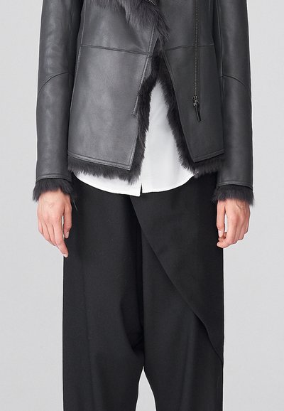 Chaqueta de cuero negra con ribetes de pelo en los bordes, cierre con cremallera y un diseño en capas, llevada sobre una camisa blanca y pantalones negros holgados.