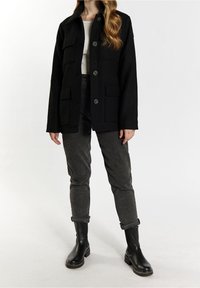 Zwarte wollen blazer met een kraag, voor knopen en vier voorzakken. Grijze, opgerolde broek en zwarte enkellaarsjes.