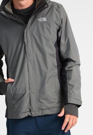 Homme portant une veste grise The North Face avec des panneaux noirs et un pantalon bleu, main partiellement dans la poche, sur fond blanc.