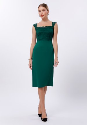 Square-neck dress featuring a lace bodice, fitted silhouette, and knee-length hem in deep green. Worn with black shoes and accessories.