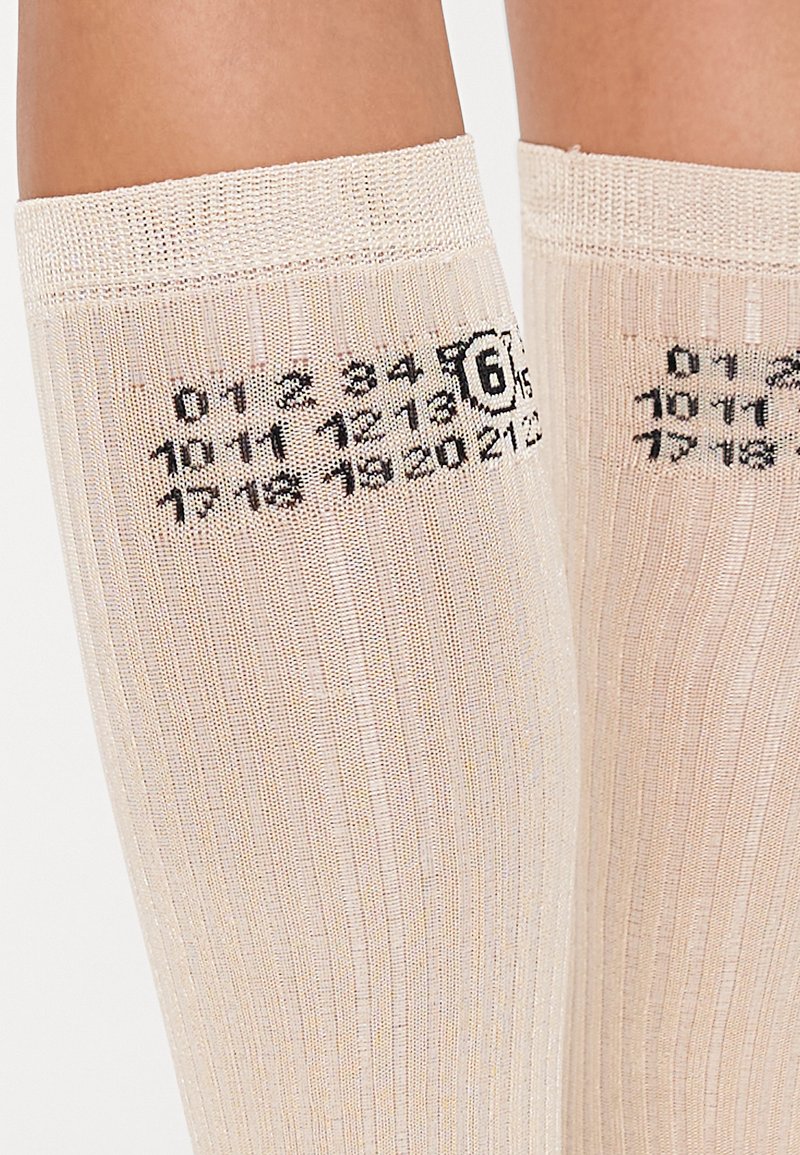 Chaussettes montantes de couleur beige clair avec une texture côtelée. Présentent des chiffres imprimés et un cercle noir avec le numéro '6'.
