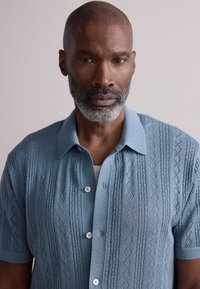 Jasnoniebieska, krótka koszulka z dzianiny z teksturowanym wzorem diamentowym, zapinana na guziki z miękkim kołnierzykiem.