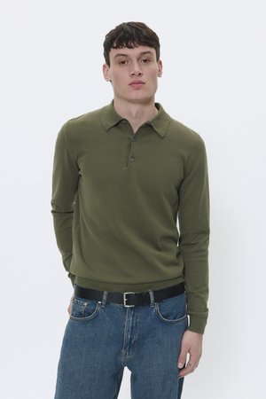 Camisa de polo verde de manga larga con cuello, que presenta una tapeta de botones y una cintura ajustada, combinada con unos jeans de mezclilla azules y un cinturón negro.