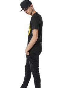 Camiseta de algodón negra con un logo amarillo en la parte delantera, combinada con jeans ajustados negros con acentos texturizados en las rodillas y una gorra negra.