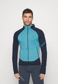 Sako na telo s tyrkysovým telom a tmavomodrými rukávmi, vyrobené z hladkej, elastickej tkaniny. Obsahuje plný zips a logo na hrudi.