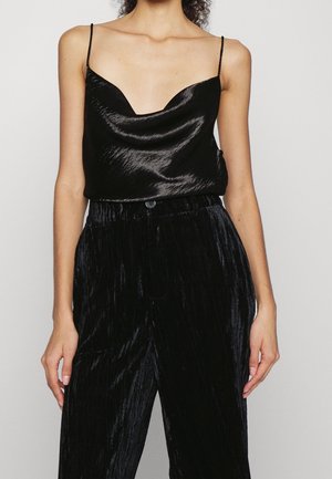 Kobieta ubrana w czarną satynową koszulkę na ramiączkach z cienkimi paskami i czarne aksamitne spodnie z wysokim stanem na jednolitym tle.