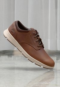 Zapatos de cuero marrón con un acabado suave, suela blanca, punta redonda y diseño clásico de cordones. Presenta un detalle de logo en el costado.