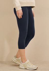 Navy blue kesim taytlar, esnek kumaştan yapılmış, açık gri detayları ve dokulu bir tabanı olan bej spor ayakkabılarla kombinlenmiş.