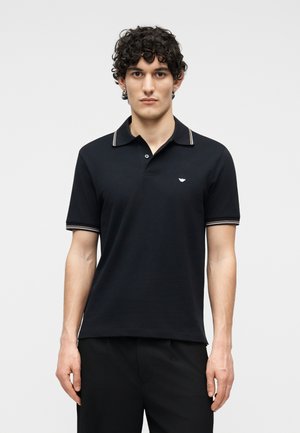 Jeune homme aux cheveux bouclés portant un polo noir à bordure blanche sur le col et les manches, se tenant devant un fond clair uni.