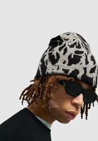 Persona con capelli ricci di castano chiaro, che indossa un cappello di lana a righe bianche e nere e occhiali da sole scuri, vestita con una maglietta nera, sullo sfondo grigio.