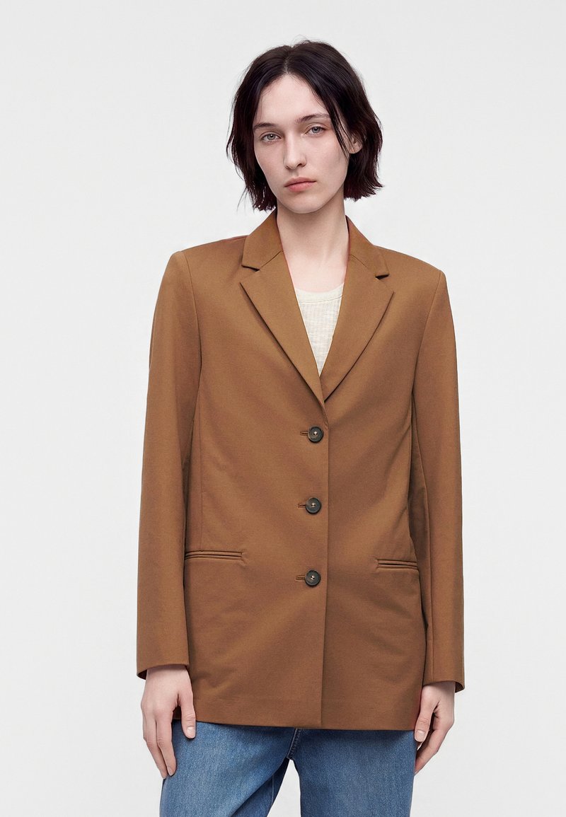 Femme aux cheveux courts et foncés portant un blazer marron à trois boutons sur une chemise claire et un jean bleu, debout devant un fond uni.