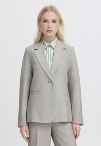 Svetlo sivi krojeni blazer s strukturiranimi rameni, en gumb za zapiranje in dvema sprednjima žepoma. Oblečen preko zelene in bele črtaste srajce.