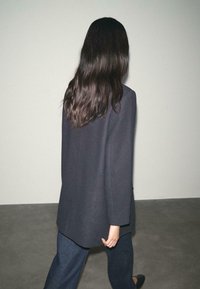 Mørk skræddersyet frakke med lange ærmer, høj krave og lige snit. Båret over brede denimbukser. Glat tekstur, minimalistisk design.