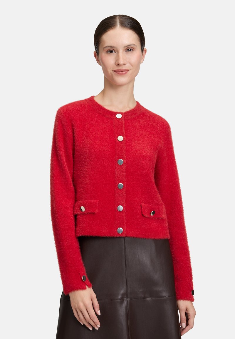 Kobieta w czerwonym, puszystym kardiganie zapinanym na guziki z klapkami, w czarnej skórzanej spódnicy, stojąca na jednolitym jasnym tle.