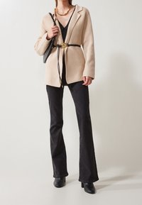 Beiger strukturierter Blazer kombiniert mit einem schwarzen Top und ausgestellten schwarzen Jeans. Accessoires sind ein Gürtel mit goldener Schnalle und eine schwarze Handtasche.