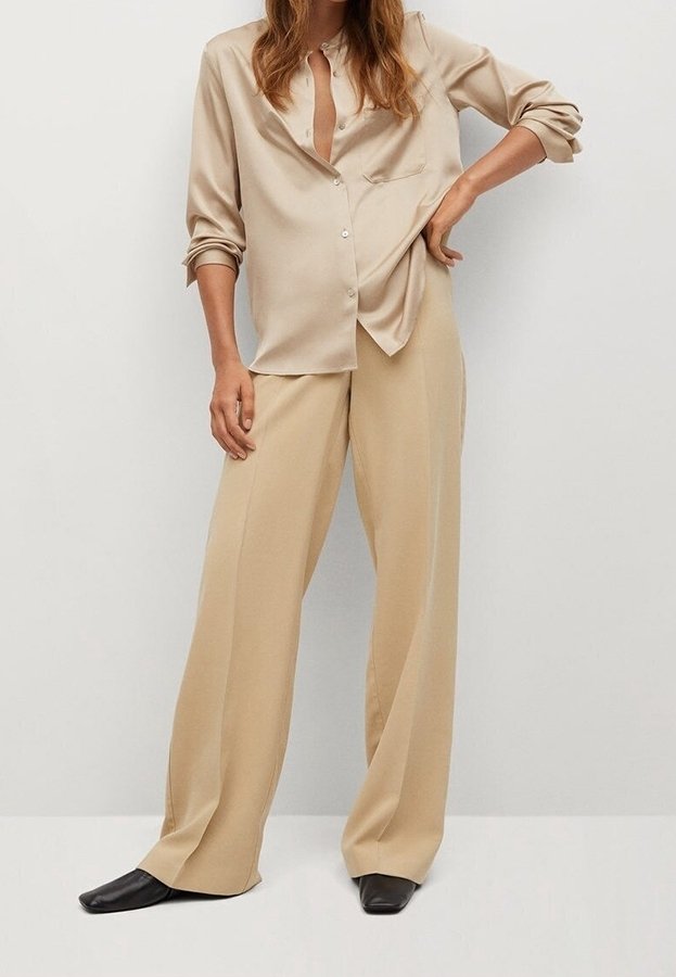 Femme portant un chemisier beige à boutons avec les manches retroussées et un pantalon large beige assorti à des chaussures noires pointues, debout devant un fond uni.