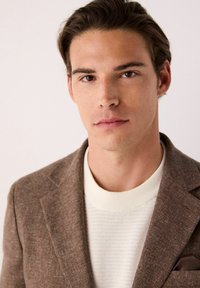 Blazer marrón con un tejido texturizado, combinado con un suéter de canalé crema. El blazer presenta un pañuelo de bolsillo en un color burdeos profundo.
