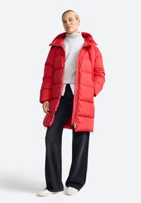 Femeie îmbrăcată într-o geacă roșie din puffer, un pulover gri cu guler înalt, pantaloni negri cu croi larg și adidași albi, stând pe un fundal simplu și deschis.