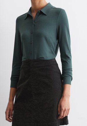 Femme portant une chemise boutonnée à manches longues vert foncé rentrée dans une jupe noire taille haute sur un fond uni.
