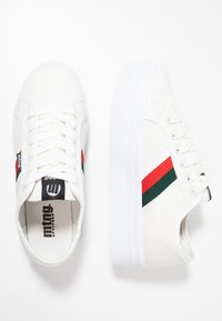 Baskets en toile blanches avec une semelle épaisse blanche, présentant des accents rayés rouges et verts et une étiquette de logo noire sur le côté.