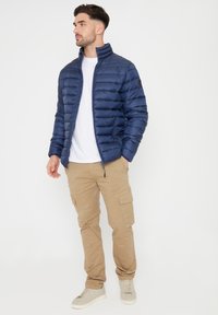 Giacca imbottita blu con colletto alto, chiusura a zip e tasche laterali, abbinata a pantaloni cargo beige e sneakers grigio chiaro.