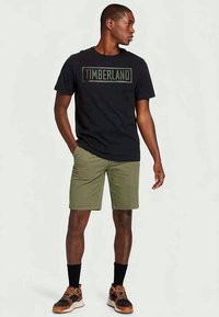 Melnas kokvilnas T-krekls ar "TIMBERLAND" drukātu pelēkā krāsā; olīvu zaļas šortas; melnas zeķes; sporta apavi ar oranžiem akcentiem.