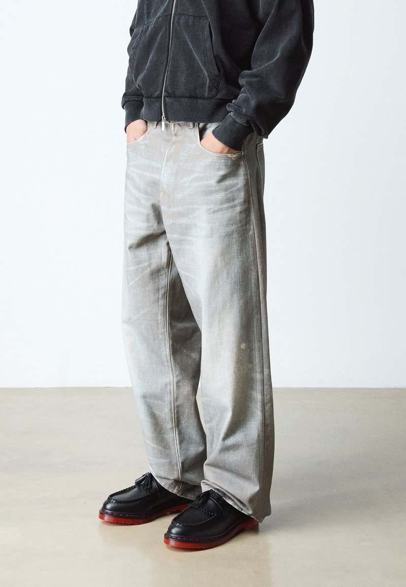 Personne portant un jean ample gris clair délavé, un sweat à capuche noir zippé et des chaussures noires aux semelles rougeâtres, debout les mains dans les poches.