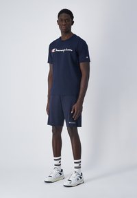 Conjunto de camiseta y pantalones cortos Champion de color azul marino, con logo en el pecho y en la pierna. Complementado con calcetines rayados en blanco, negro y rojo, y zapatillas deportivas.