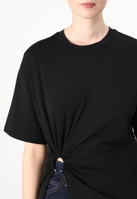 Tricou negru din bumbac, cu mâneci scurte, detaliu cu nod strâns în față și o deschidere circulară, purtat peste blugi de culoare albastru închis.