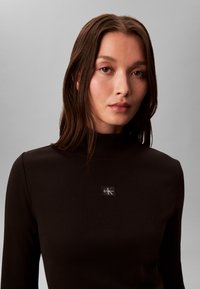 Femme aux cheveux bruns mi-longs portant un haut à col roulé côtelé noir avec un logo Calvin Klein sur la poitrine.