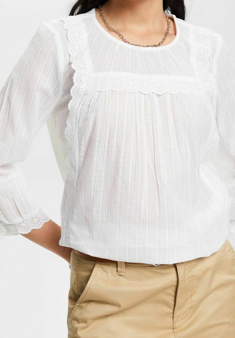 Femme portant un chemisier blanc bordé de dentelle avec des manches bouffantes et un pantalon beige, debout, une main sur la hanche.