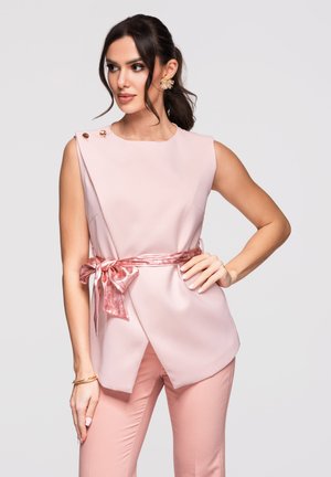 Vrouw draagt een mouwloos lichtroze topje met decoratieve knopen en een roze zijden sjaalriem, gecombineerd met bijpassende roze broek.