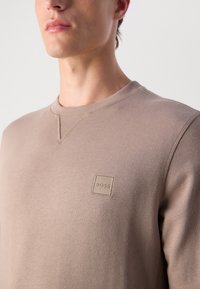 Sweatshirt beige en tissu doux, avec un col rond et un petit patch carré en logo avec "BOSS" brodé au centre.