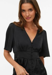 Sort kjole laget av glatt stoff, med korte ermer, V-hals, knapper foran og en detaljert midje med rynking.