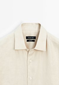Camicia di lino beige con colletto alla francese, cinque bottoni e una consistenza liscia. Etichetta di cura visibile all'interno del colletto.