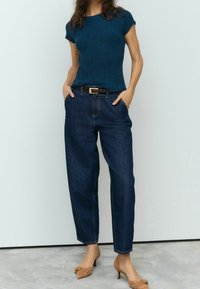 Mørkeblå højtaljede jeans med en afslappet pasform, parret med en tætsiddende teal ribstrik top og lysebrune hæle med sløjfedetalje.