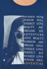 Granatowa koszulka z grafiką czarno-białą przedstawiającą kobietę w okularach przeciwsłonecznych, z białymi i żółtymi napisami podkreślającymi słowa związane z wzmocnieniem.