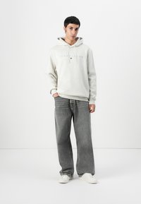 Lys grå hettegenser med "TOMMY JEANS" tekst, kombinert med løse, mørkegrå jeans. Skoene er hvite joggesko. Enkel, uformell design.