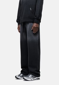 Zwarte hoodie met een voorzak, gecombineerd met donkergrijze joggingbroek met een trekkoord. Schoenen zijn zilver en zwarte hardloopsneakers.