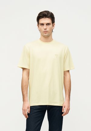 Homme portant un t-shirt à manches courtes jaune pâle et un jean foncé, debout devant un fond clair uni, regardant droit devant.