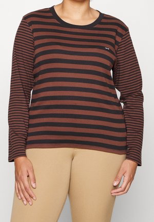 Persoon die een lange mouwen zwart en bruin horizontaal gestreept shirt draagt met beige broek, staande tegen een effen achtergrond.