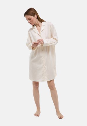 Femme portant une robe-chemise ample à manches longues couleur crème, pieds nus, debout devant un fond clair uni, regardant vers le bas.