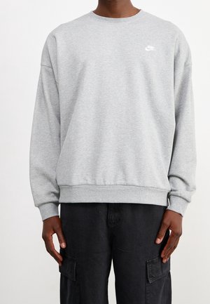 Personne portant un sweat-shirt Nike gris clair à manches longues et un pantalon noir, debout devant un fond blanc uni.