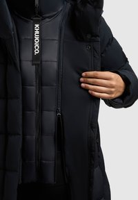 Hånd justerer lynlåsen på en sort, polstret vinterjakke med synlige indre og ydre lag samt en mærket trækflig.