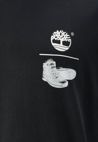 Black cotton t-shirt featuring a white graphic of a tree logo above a pair of sneakers. The design is centered on the upper front.