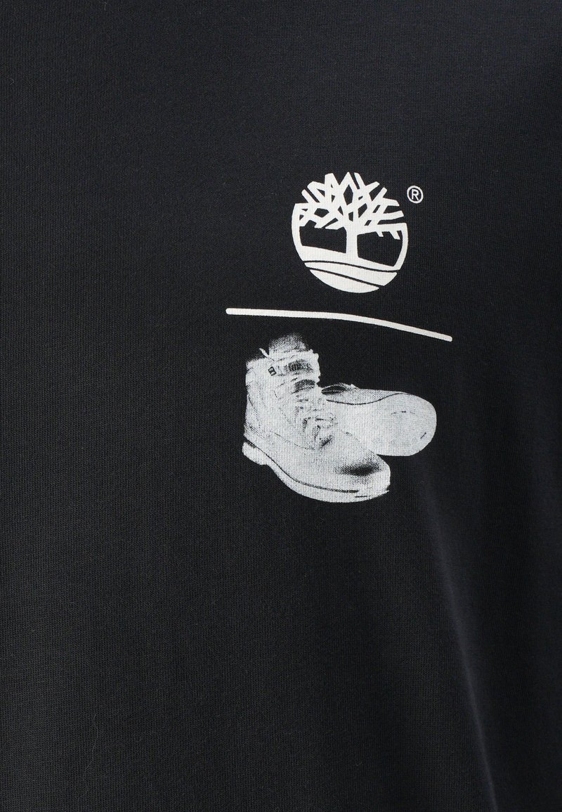 T-shirt in cotone nero con un grafico bianco di un logo ad albero sopra un paio di sneakers. Il design è centrato sulla parte superiore frontale.