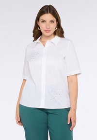 Camicetta bianca a maniche corte con bottoni, decorata con scintillanti ornamenti blu, gialli e verdi sul lato sinistro, abbinata a pantaloni teal.