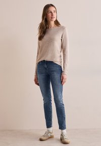 Pull à manches longues beige avec un col rond, associé à un jean bleu slim et des baskets beiges. Texture simple, coupe décontractée et sans motifs discernables.
