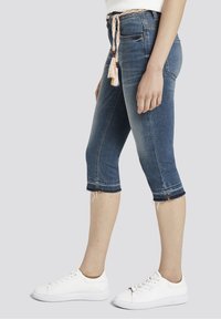 Pantalones capri de mezclilla en tono oscuro con dobladillo deshilachado, que presentan un cinturón trenzado y bolsillos laterales, combinados con zapatillas blancas.
